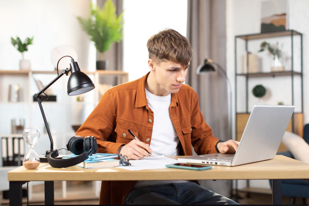 Focused young man in casual wear sitting at table with lamp and modern laptop and making notes. Concept of people, technology and distance learning.