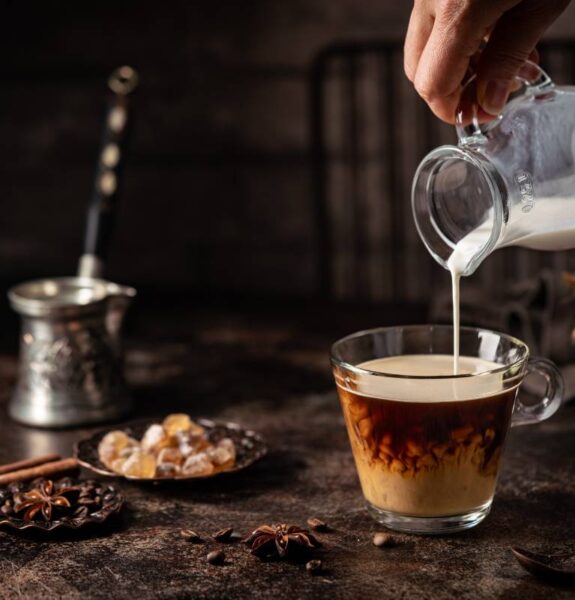 Coffee in a glass with cream poured over, sugar and coffee beans on dark background