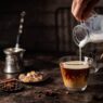 Coffee in a glass with cream poured over, sugar and coffee beans on dark background