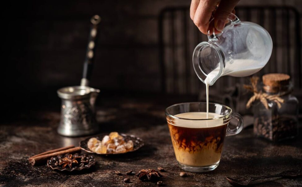 Coffee in a glass with cream poured over, sugar and coffee beans on dark background